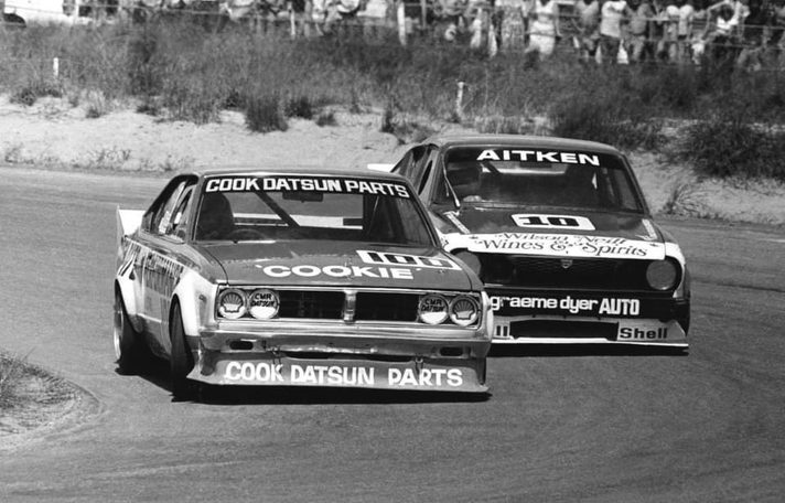 Reg Cook's Datsun #100 leads Grant Aitken's #10 through a sandy corner, spectators behind