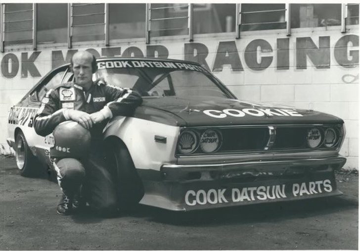 Reg Cook in his racing overalls, helmet in hand, kneeling beside the Cook Datsun Parts car 