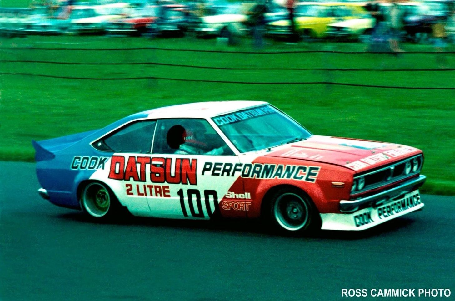 The Cook Datsun 160J #100 in colour, red bonnet and blue tail, at speed past a crowd