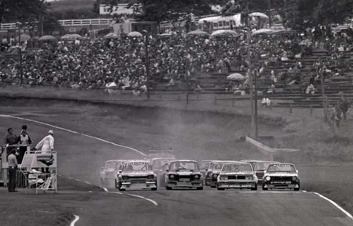 A saloon field starts in front of a packed grandstand, the Cook Datsun and Aitken's #10 on the front row